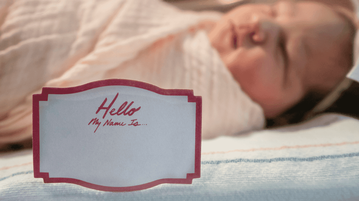 A baby in a hospital cradle with a blank name tag on it saying "Hello My Name Is"