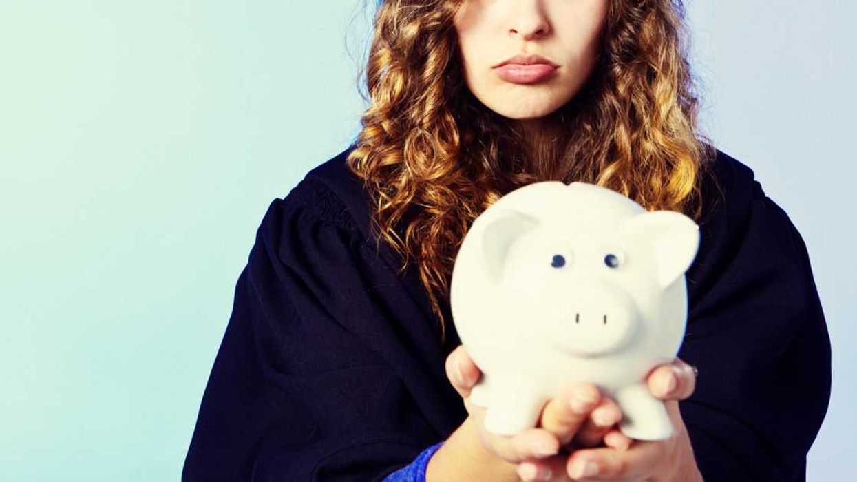 A beautiful young honey-blonde woman in academic dress looks disappointed and she is pouting as she holds up a piggy bank.