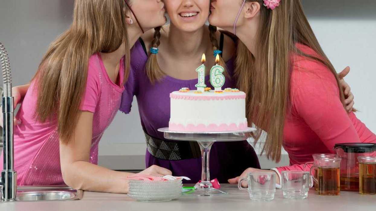 A birthday girl is kissed on both of her cheeks by two other girls in front of her birthday cake with one and six candles