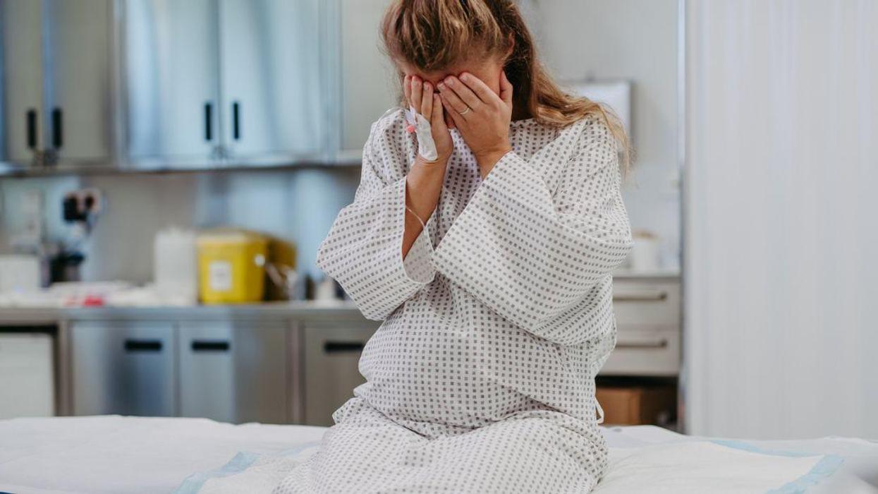A blonde, pregnant woman sits on an examination table, sad with her face in her hands.