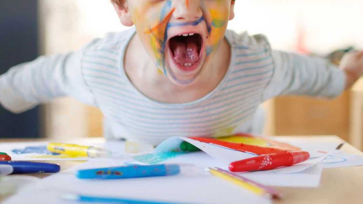 A boy paints his face with colorful felt pens, smiles and stares into the camera.