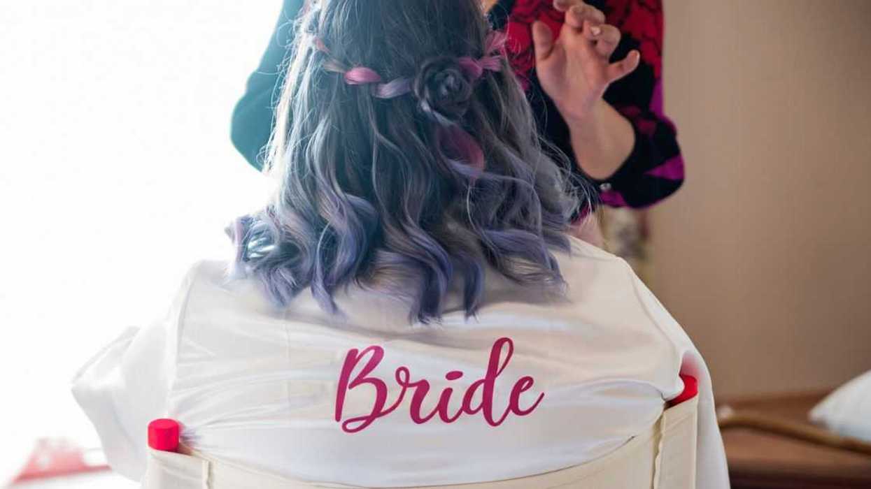A bride gets her make-up done. She is not facing the camera, the back of her robe reads bride in red letters.