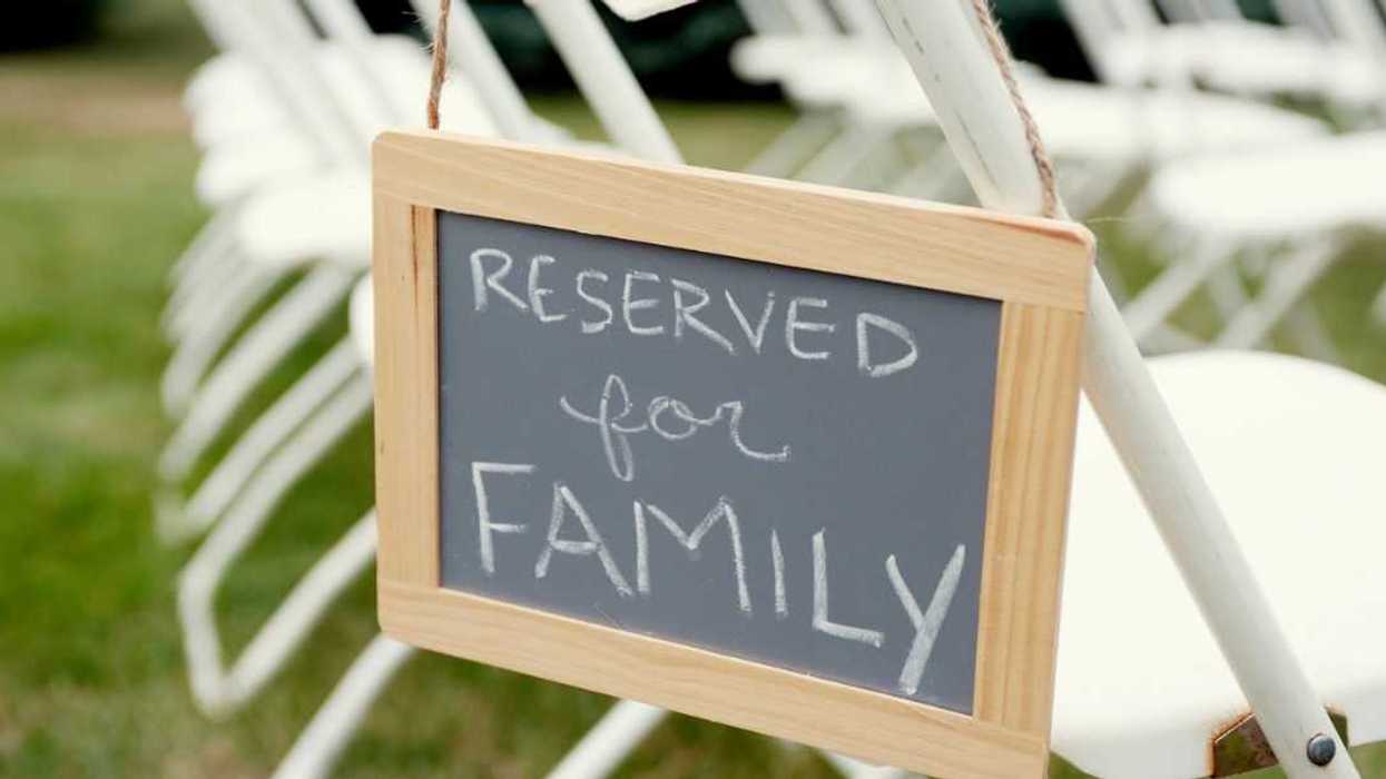 a chalkboard board signs hangs off of a white chair at a wedding. It says,