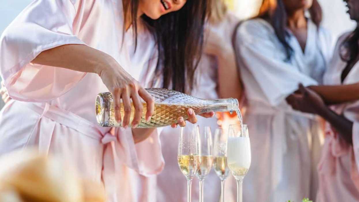 A cheerful bride in a robe pours champagne into glasses at a bachelorette party with friends in pastel robes, celebrating outdoors.
