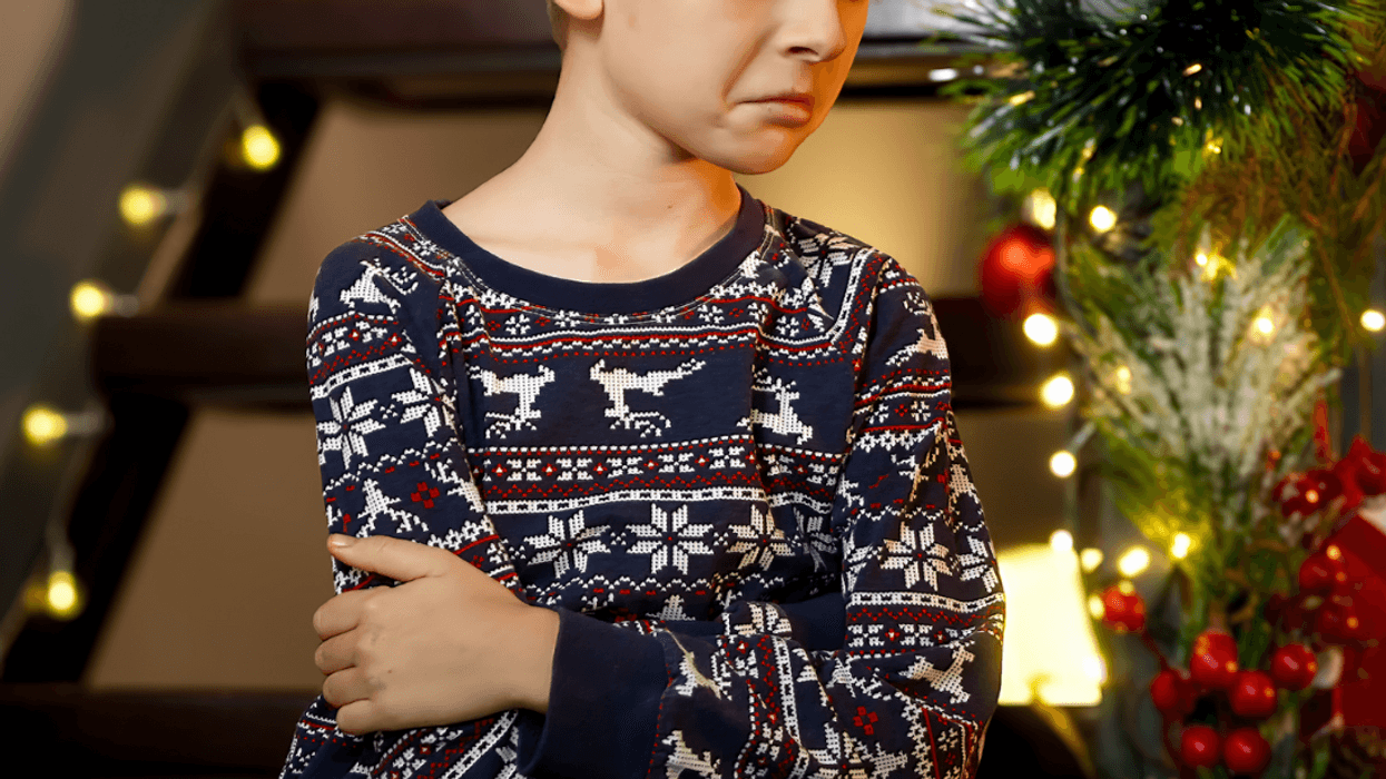 A child with a frown on his face, folding his arms, standing in front of a Christmas tree.