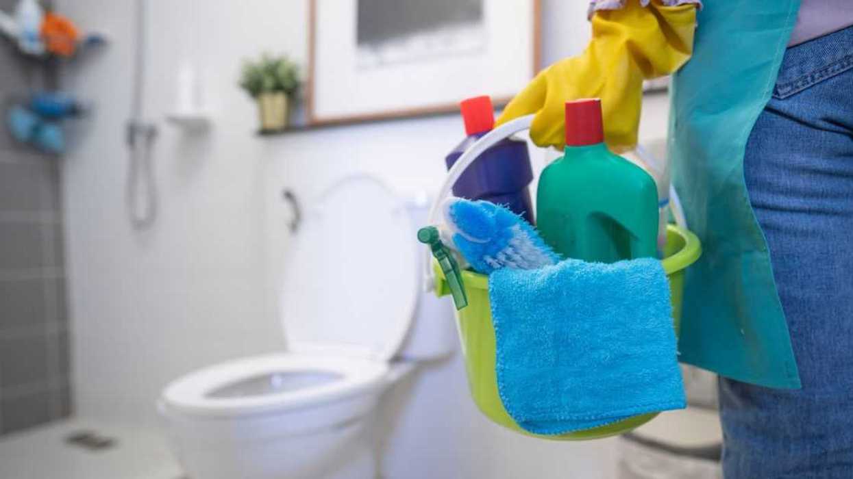 A cleaning woman is standing in the bathroom holding a blue bucket full of chemicals and facilities. The toilet is in the corner.