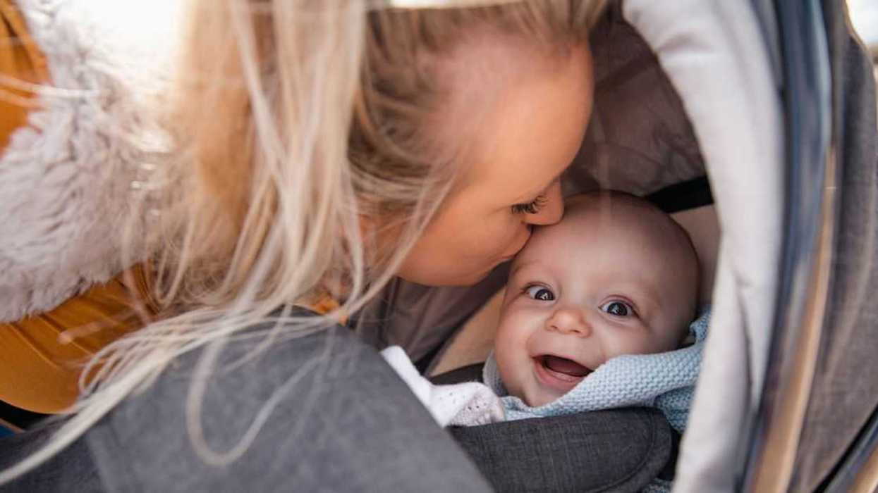 A close up shot of a mother kissing her son on the head, the baby boy is in his stroller and the woman is crouching down next to him.