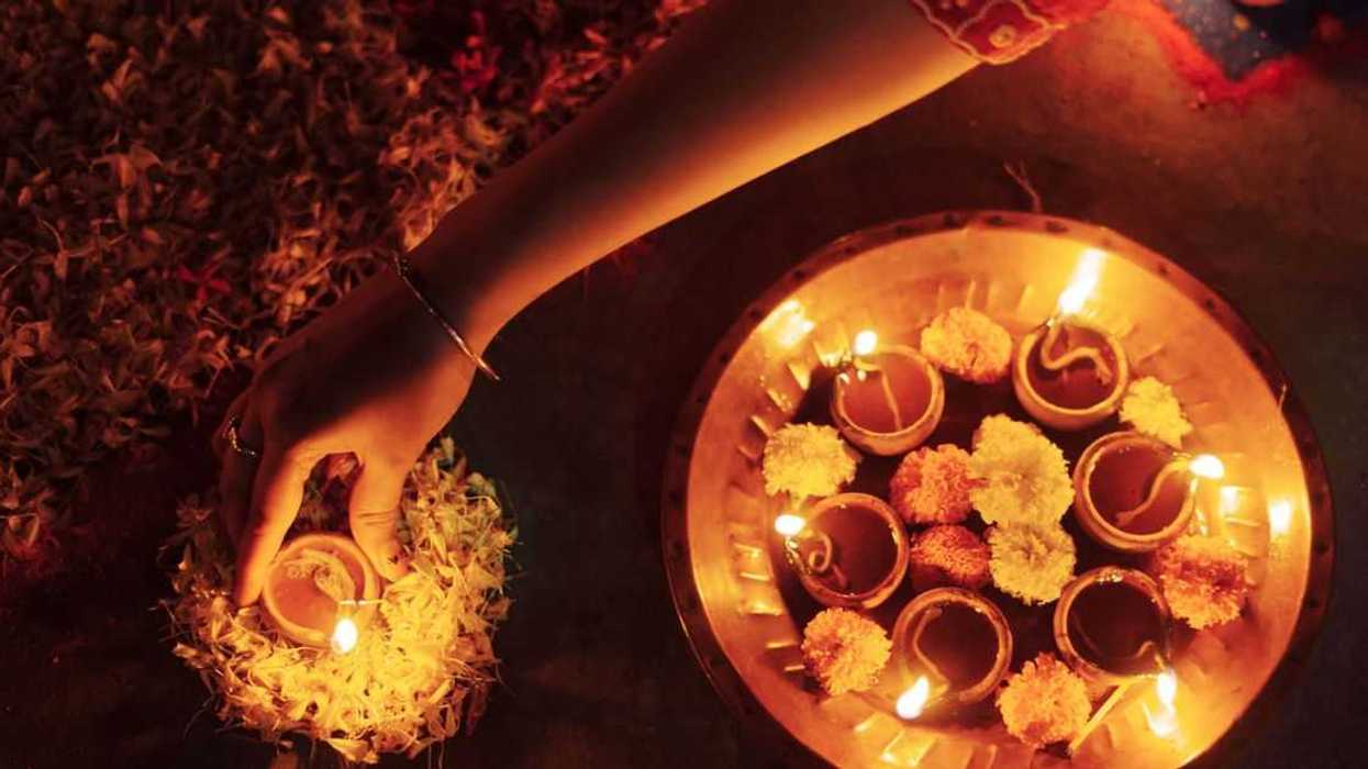 A close-up view of a woman’s hand placing a lit clay lamp (diya) surrounded by marigold flowers, creating a warm and serene ambiance during Diwali, the Hindu festival of lights. This traditional ritual symbolizes the victory of light over darkness.