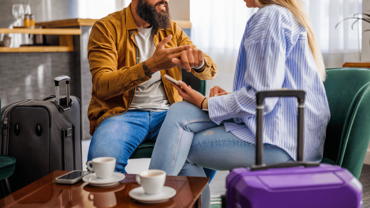 A couple sitting at a table talking to each other at a table with cups of coffee on it and luggage in front of them.