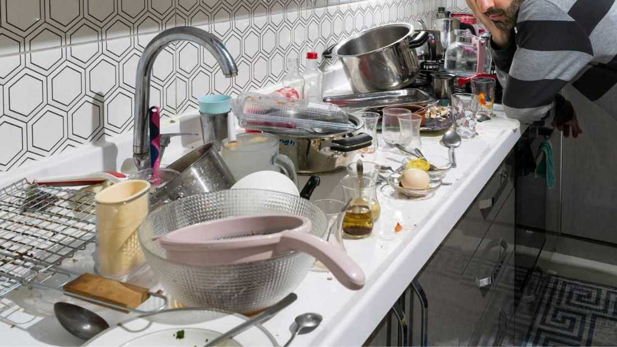 A depressed looking man stares at a sea of dirty dishes strewn along a kitchen counter.