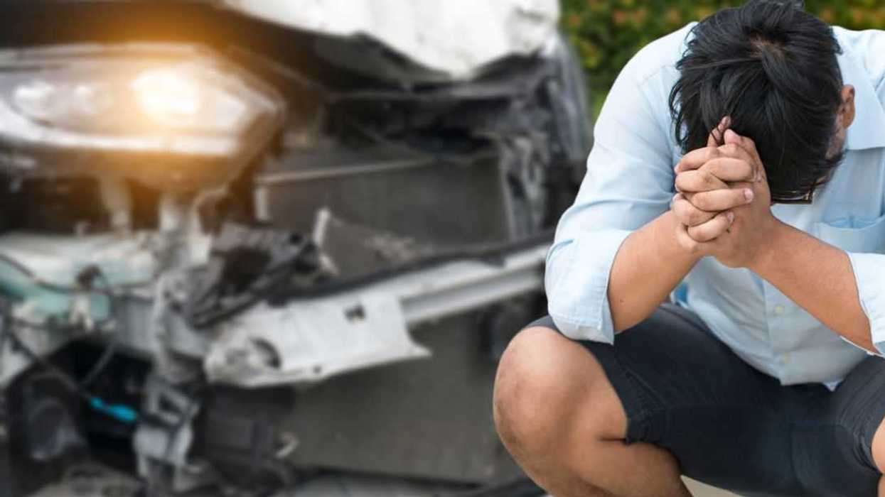 A distressed man sits on the curb in front of a car accident, he has his head in hands.