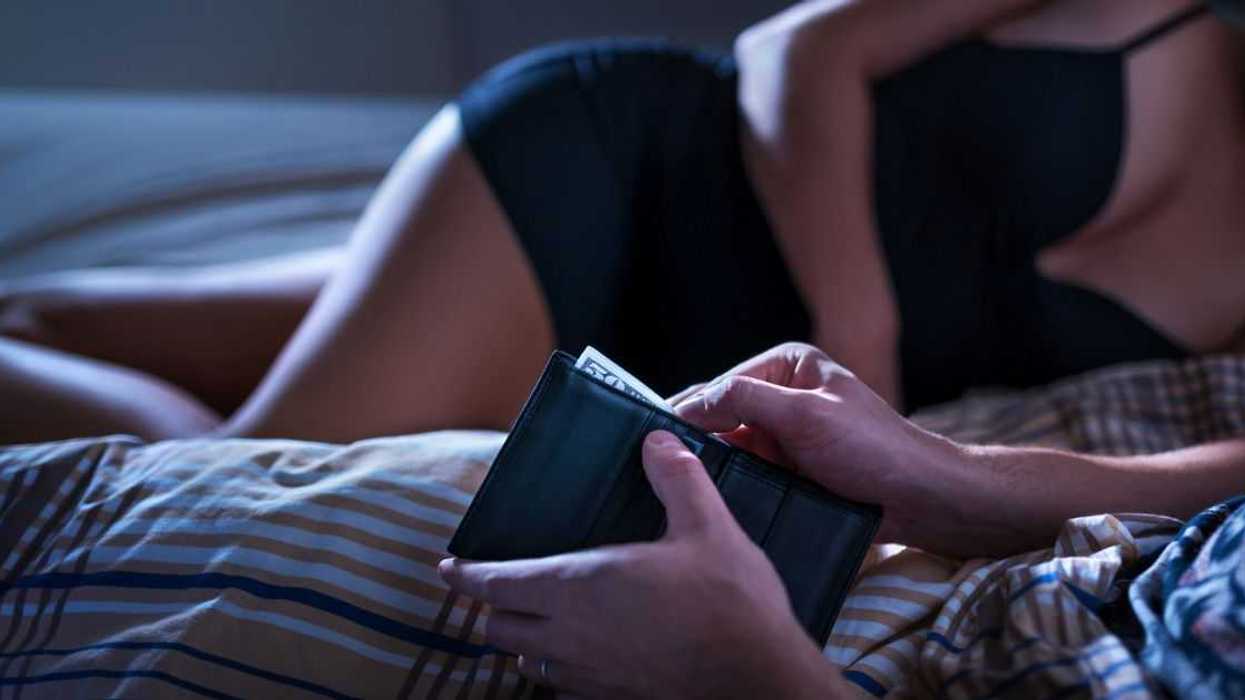 A female escort and man lay in bed as he retrieves money from his wallet