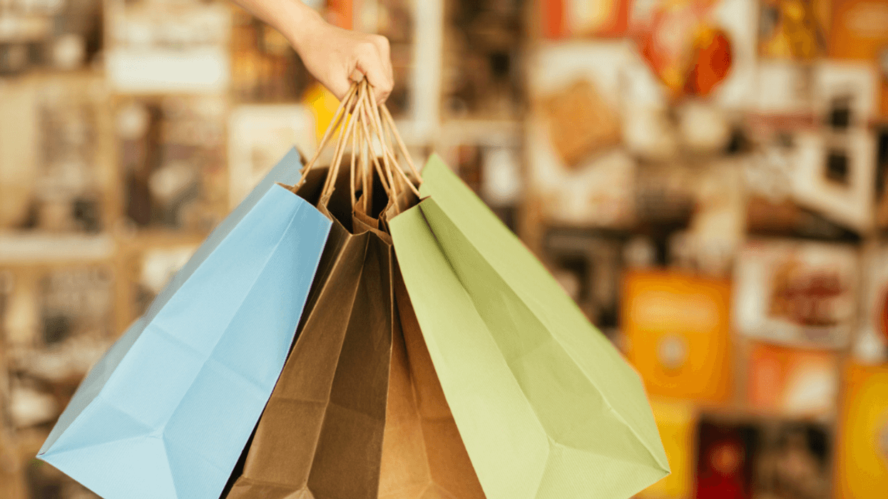 A hand holding three different colored paper shopping bags.
