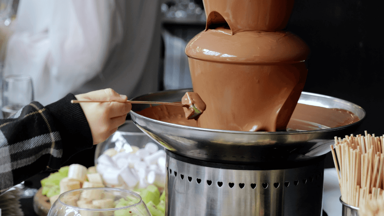 A hand sticking a piece of fruit into a chocolate fountain.