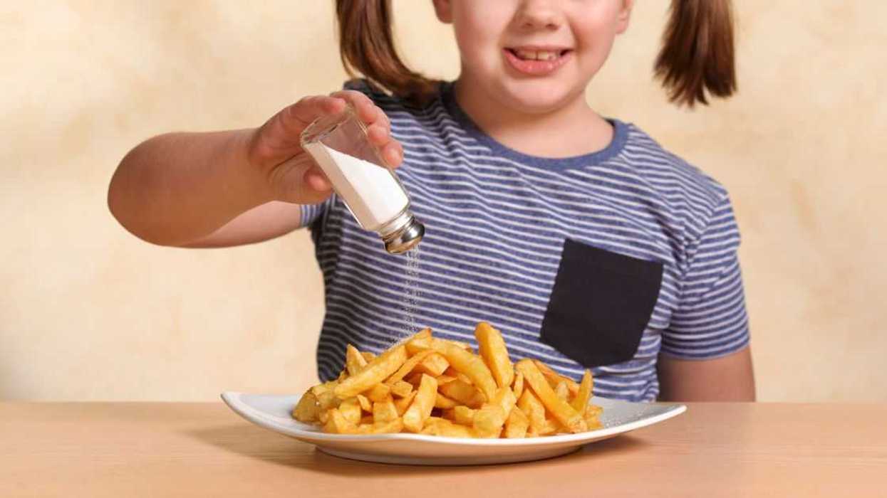 A little girl sits at a table and shakes salt on her fries.