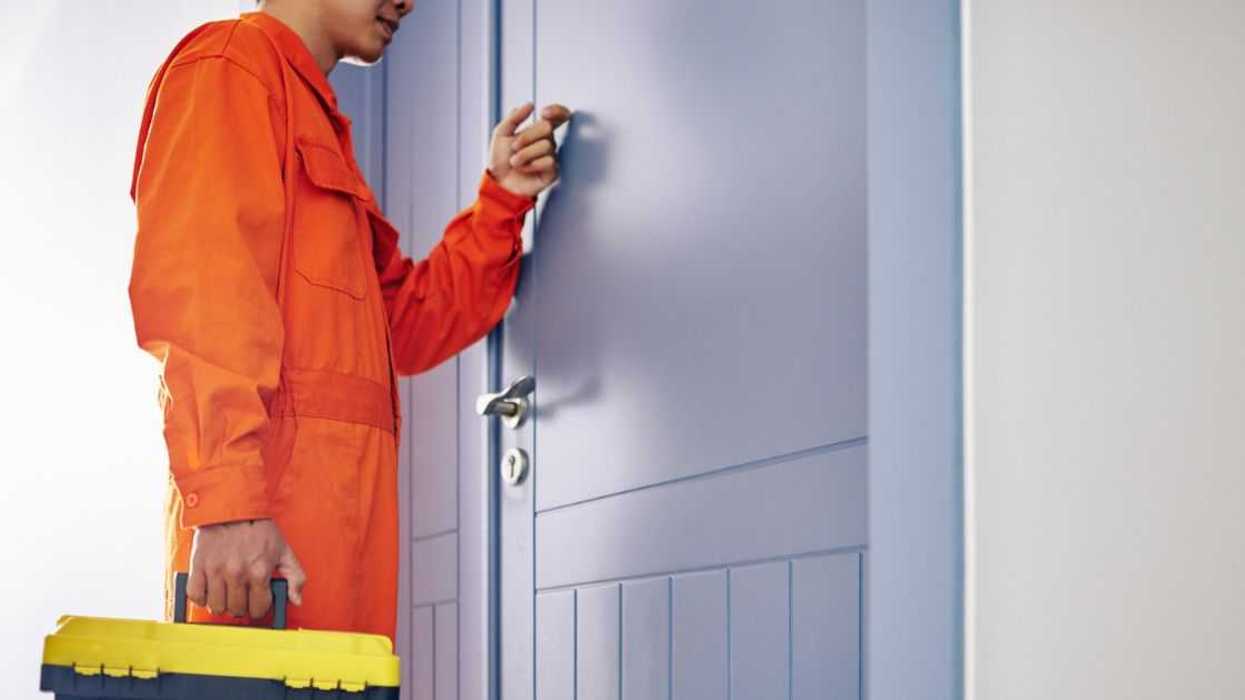 A maintenance man dressed in orange overalls knocks on a door