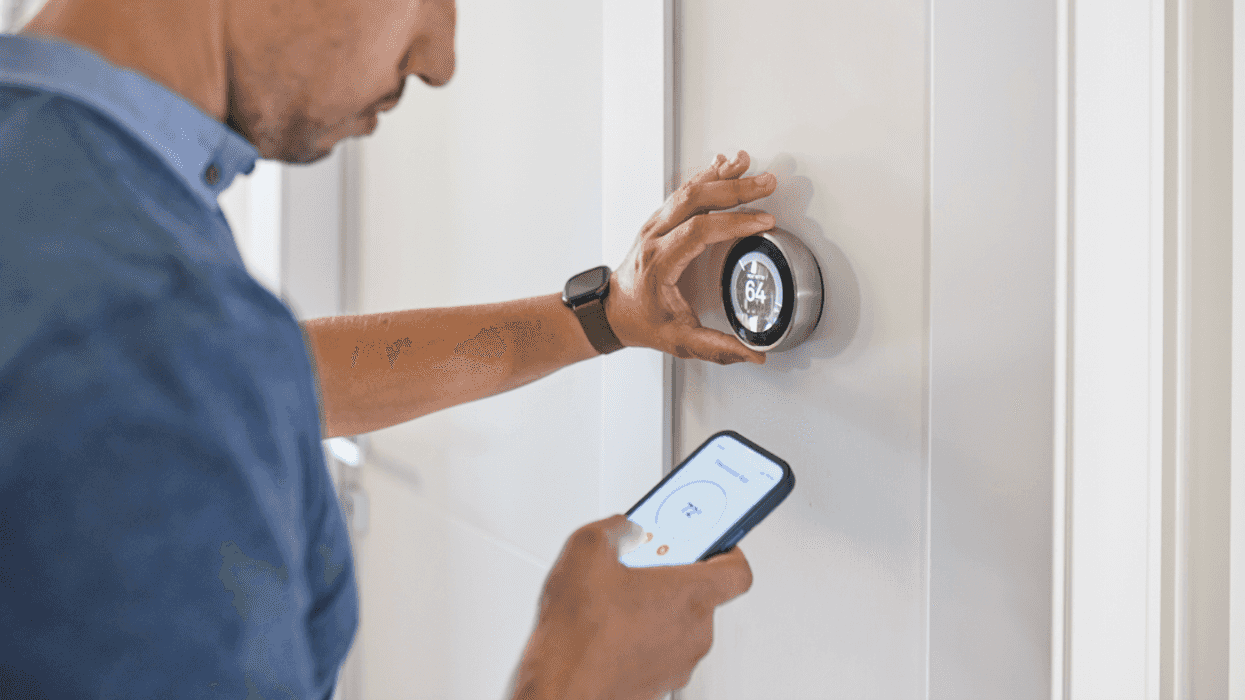 A man adjusting a thermostat on the wall and with his phone.
