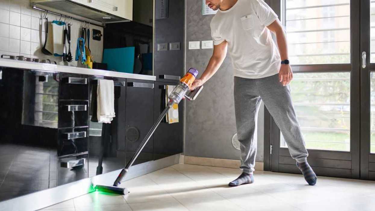 A man cleaning the floor in a modern kitchen and living area using a cordless vacuum cleaner. The room is bright with natural light, featuring a contemporary design with reflective black cabinets, a wall calendar, and large windows. The man is casually dressed and focused on cleaning, with the vacuum's green laser light illuminating the floor.