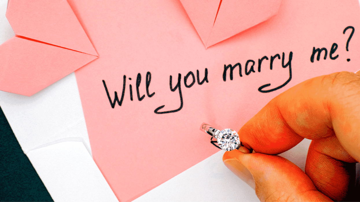 A man holding a ring in front of a pink piece of paper with "will you marry me" written on it.