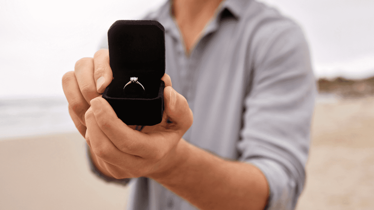 A man holding out an open ring box with a diamond ring inside it.
