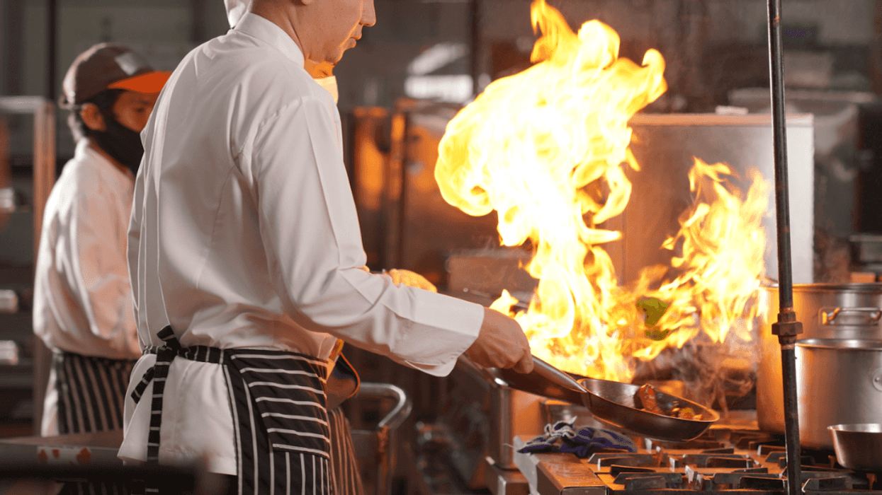 A man in a chef's coat in front of a stovetop with a big flame coming out of a frying pan.