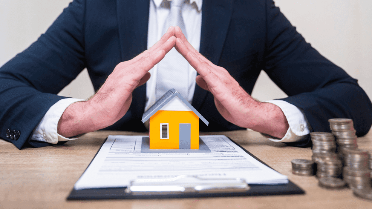 A man in a suit sitting at a desk with his fingers touching over a contract and a miniature house.