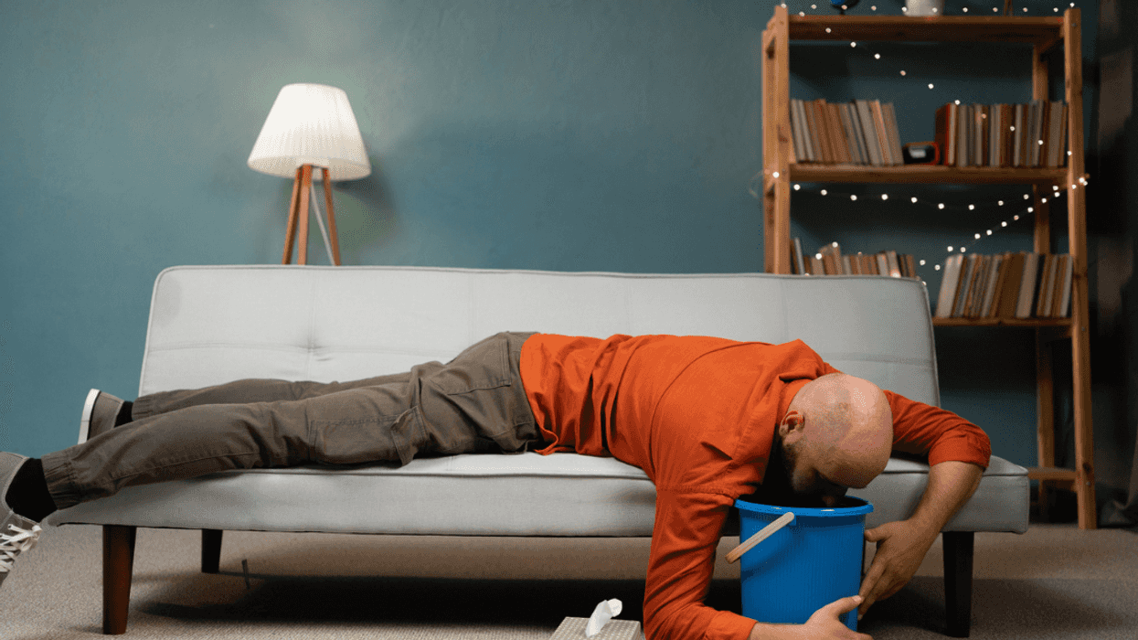 A man lying on his stomach on the sofa and throwing up into a bucket.