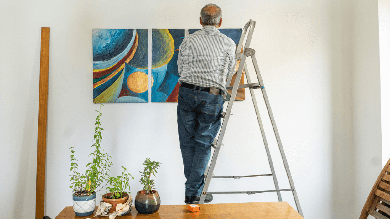 A man on a ladder hanging a painting on the wall