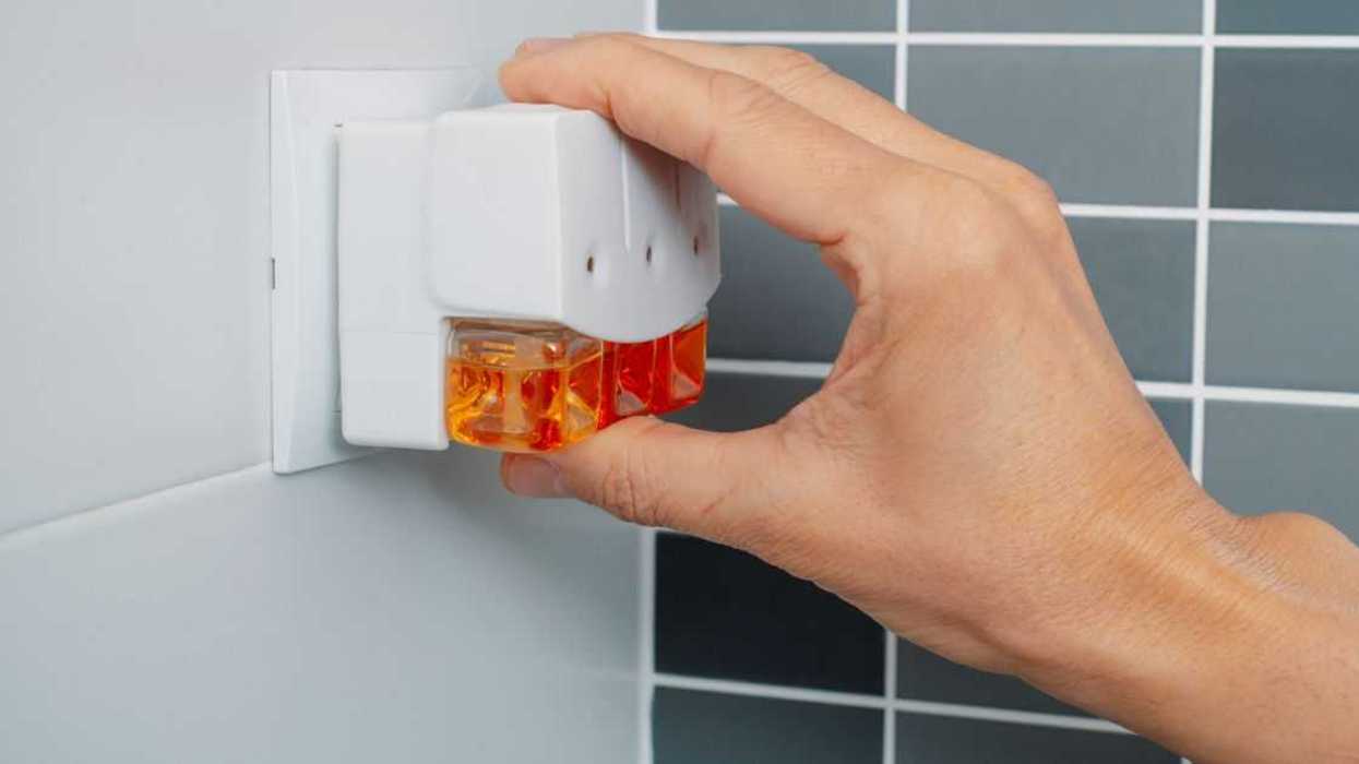 A man's hand plugs a plug-in air freshener into a socket on a tiled bathroom wall.