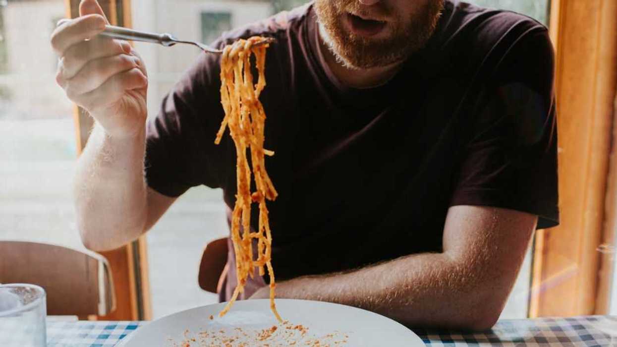 A man sits at a table, covered with a blue checkered tablecloth. He uses a fork to scoop up a mouthful of long spaghetti pasta. Meatballs rest on the large plate.
