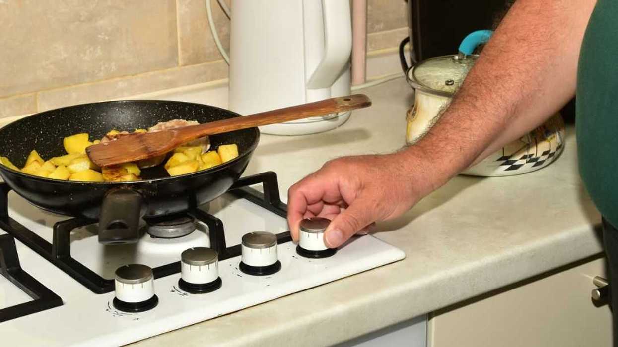 A man turns the knob of a gas stove to turn off the heat on the burner underneath a frying pan.