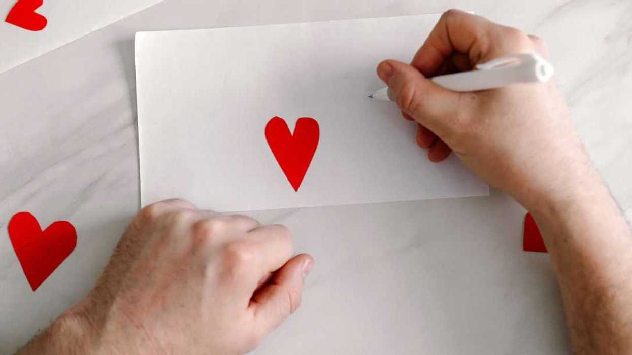 A man writes a heartfelt note saying I love you on an envelope, surrounded by red hearts and a single rose on a marble table, creating a romantic atmosphere.