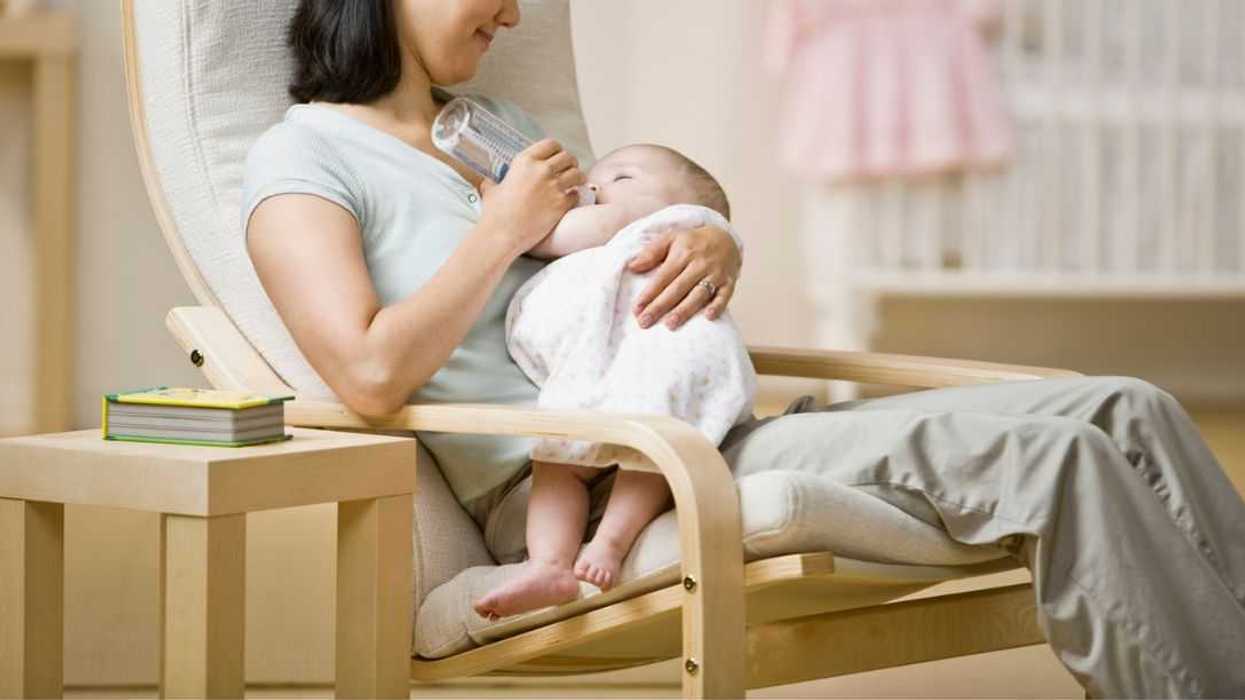 A mother sits in a chair while bottle feeding a baby.