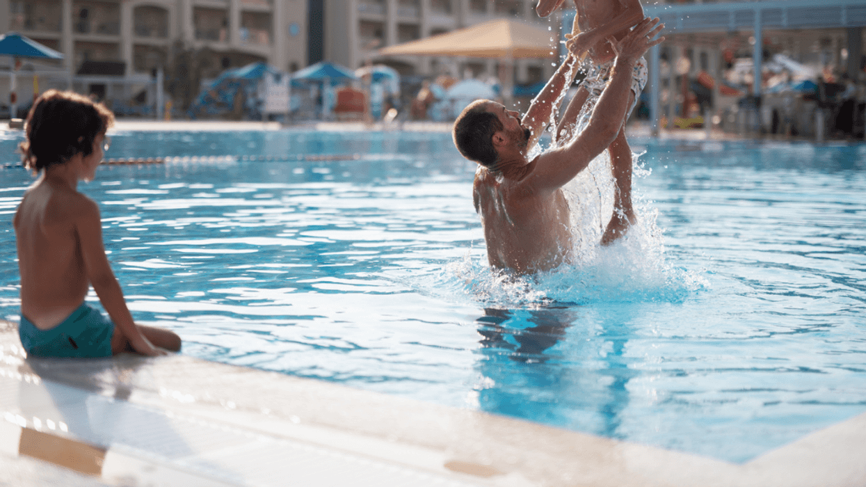 A pan throwing a child in the air in a pool with another child looking on.