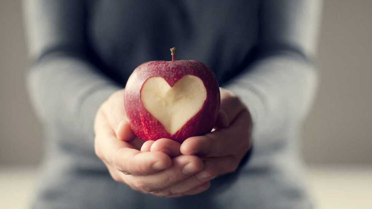 A person holds a red apple in their hands. There is a heart carved out of the apple.