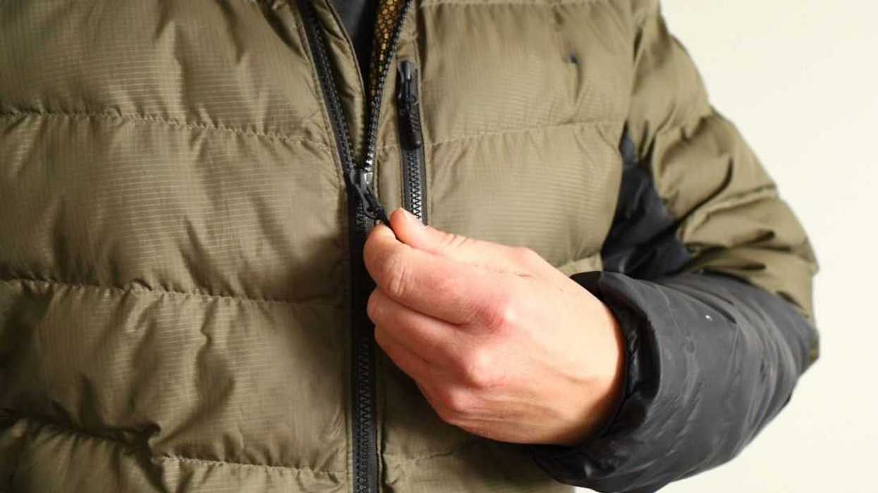 A person in a green puffer jacket zipping up, focus is on his hand and zipper, neutral background.