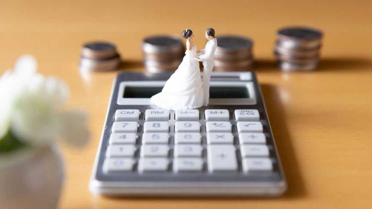A plastic figure of a married couple sits on top of a calculator. Stacks of coins sit behind them against a yellow background.