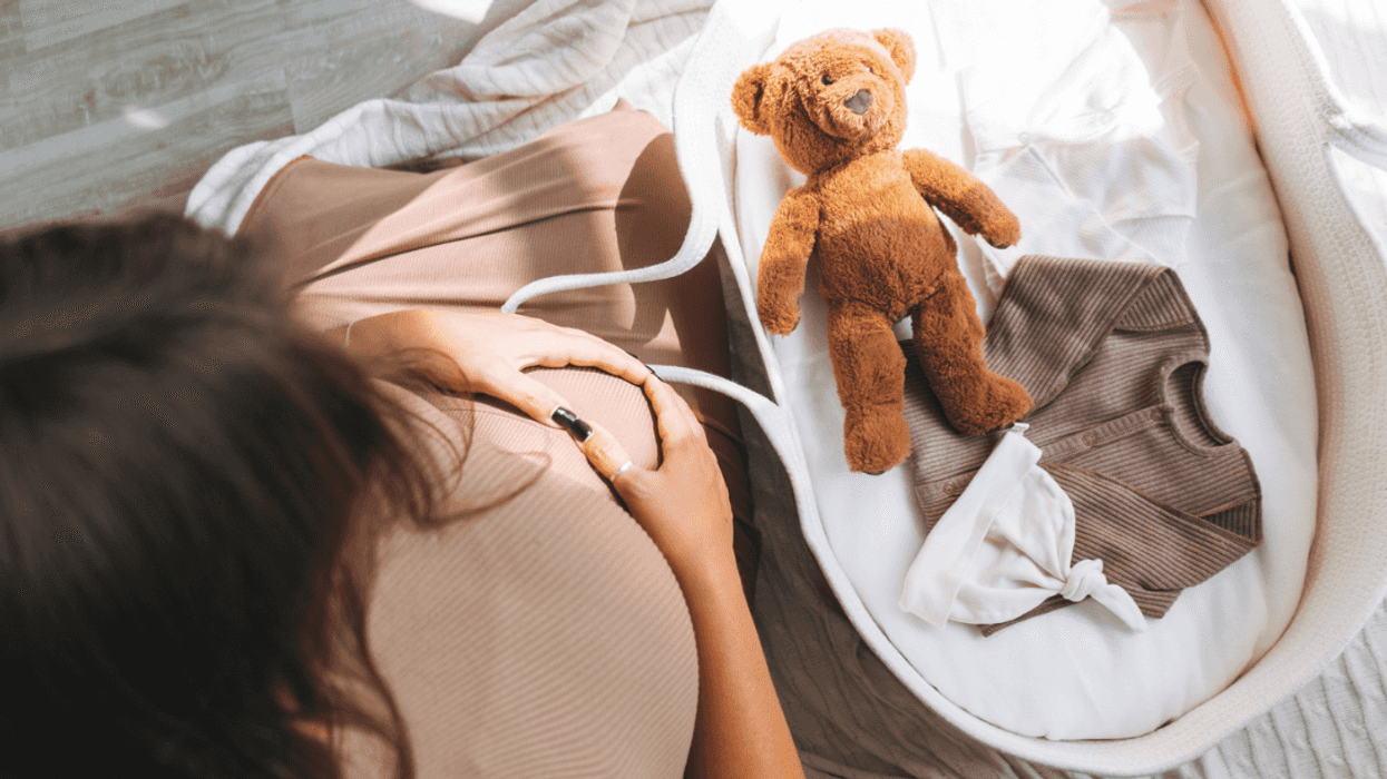 A pregnant woman looking down over a crib with a teddy bear in it.