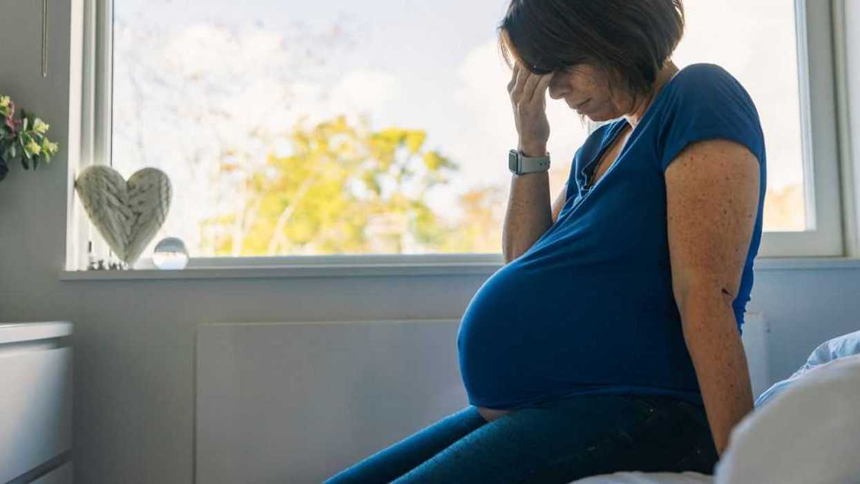 A pregnant woman sits on the edge of a bed seemingly in despair