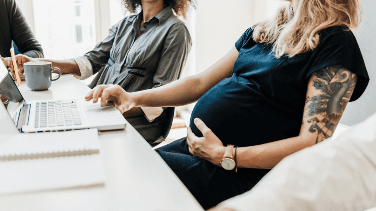 A pregnant woman sitting at a computer, talking to another woman at the same desk.