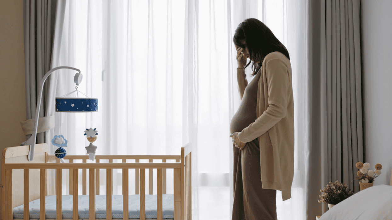 A pregnant woman standing and crying over a crib.