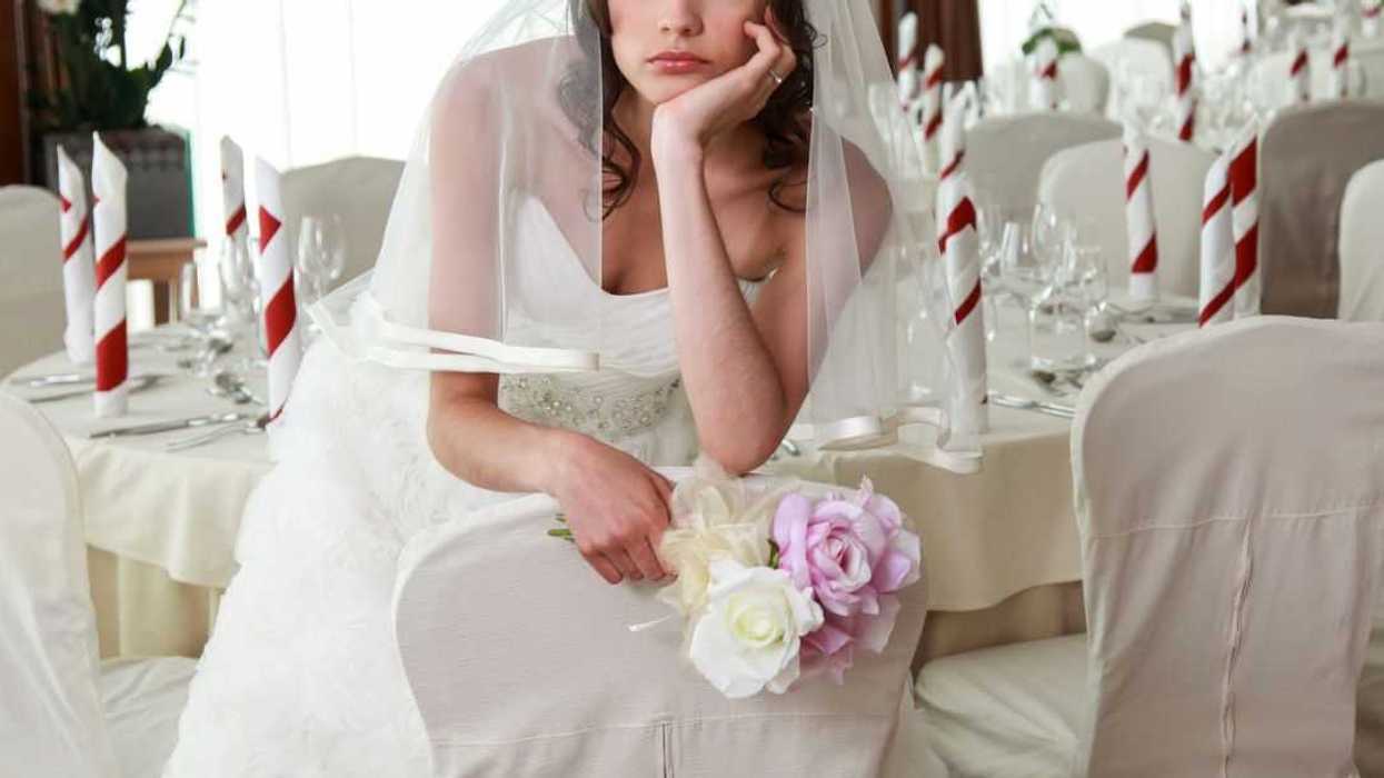 A sad bride sits in an empty banquet room prepared for a wedding, she holds flowers.