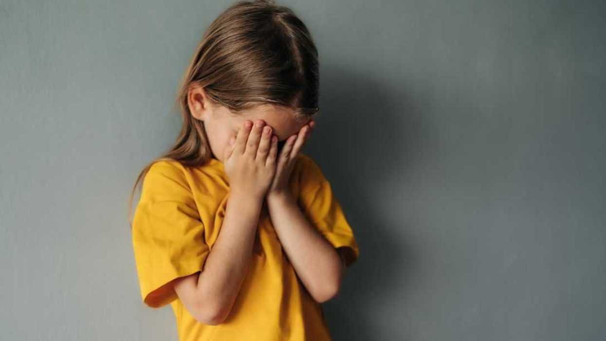 A sad, young girl cries into her hands, she stands against a gray backdrop.