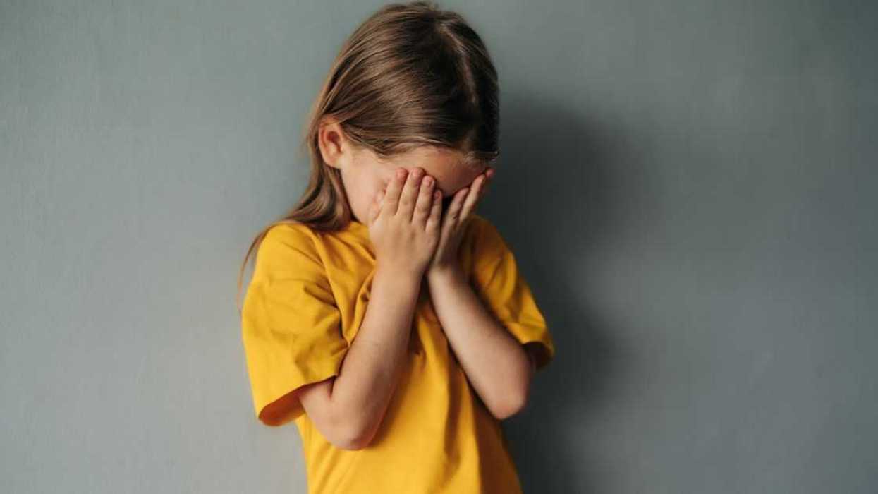 A sad, young girl cries into her hands. She wears a yellow shirt and stands in front of a gray background.