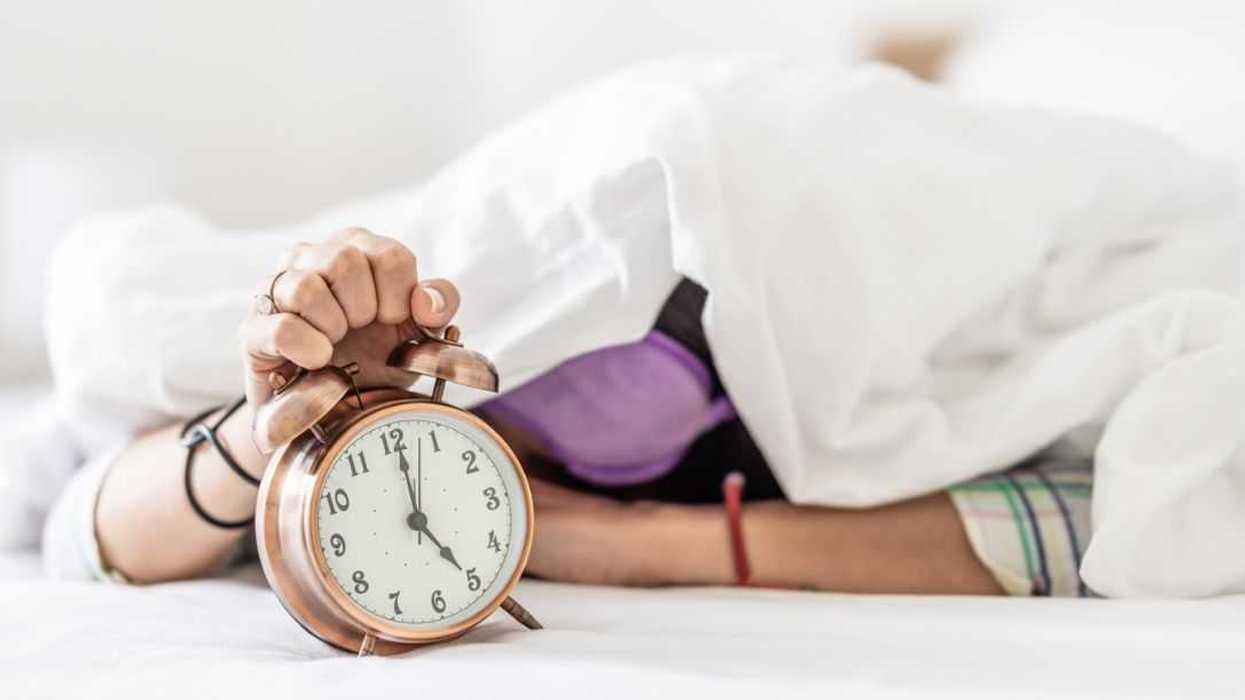A sleepy young woman hides under the duvet as she is awakened by the loud sound of the alarm clock.
