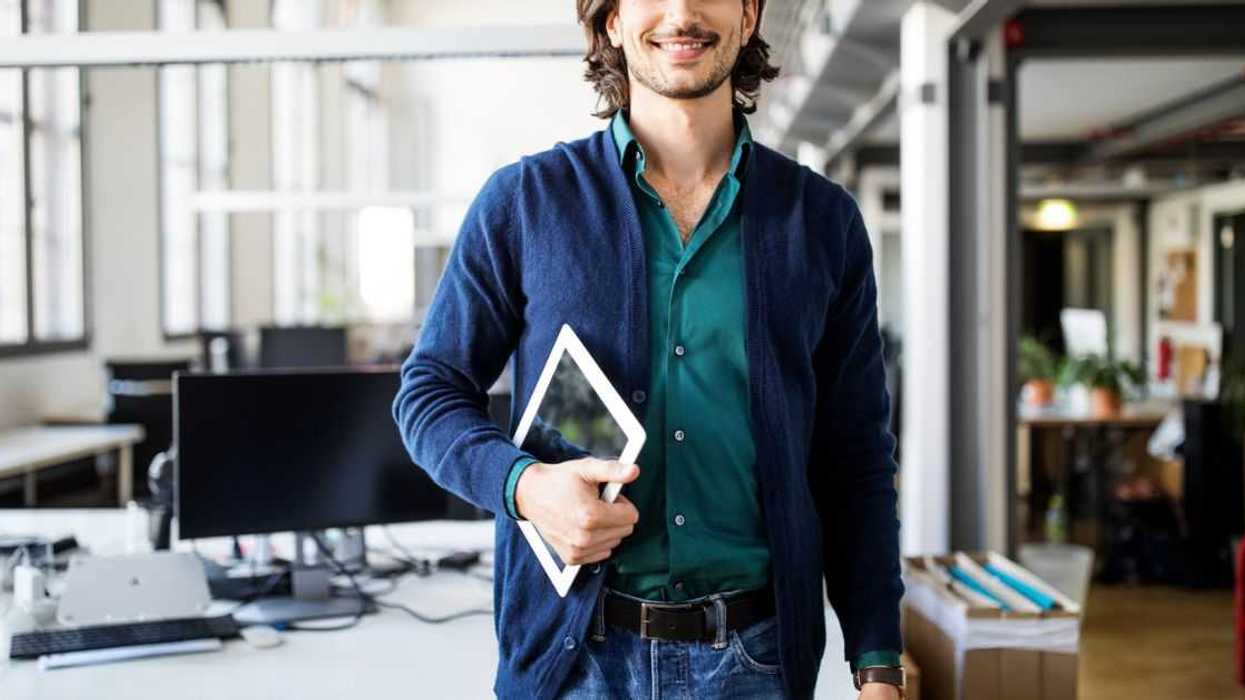 A smiling young man with should length hair is dressed casually, holding a tablet