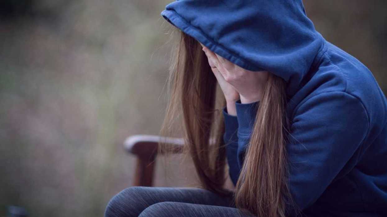 A teenage girl in hooded top, with head in hands in despair. She sits on a park bench.