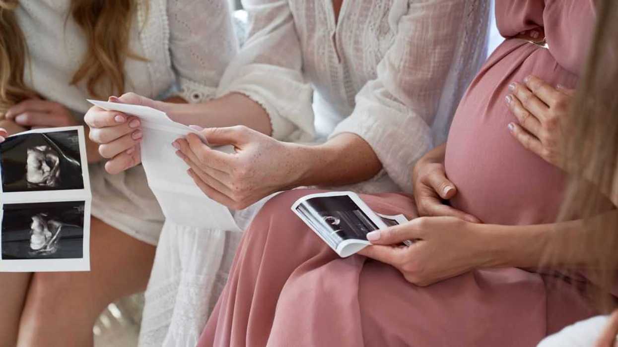 A very pregnant woman is surrounded by women at a baby shower as they look at sonograms