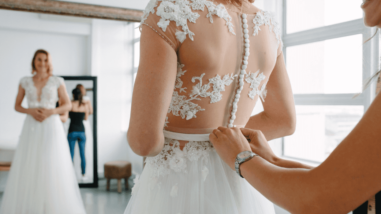 A woman buttoning up a woman in a wedding dress from behind.