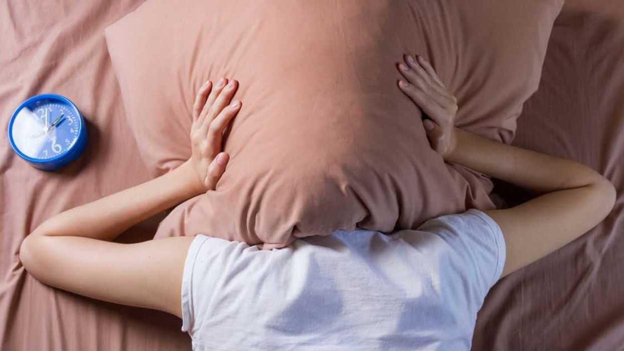 A woman covers her head with a pillow. An alarm clock sits next to her.